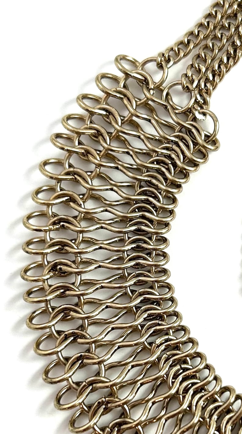May include: A gold-toned metal chain necklace with a unique, woven design. The chain is made of multiple interconnected loops, creating a textured and intricate pattern.