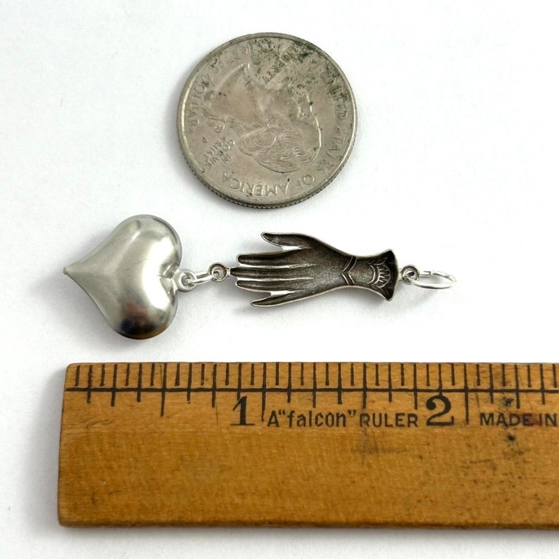 Puede incluir: Collar con dije de plata con un coraz&oacute;n y un dije de mano. Los dijes miden aproximadamente 2,5 cm de largo. El collar se muestra junto a una regla para comparar el tama&ntilde;o.