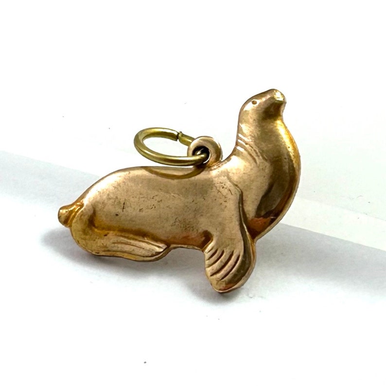 May include: A gold-toned charm in the shape of a seal. The charm has a loop at the top for attaching to a necklace or bracelet.