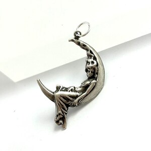 May include: A silver charm pendant featuring a woman sitting on a crescent moon.