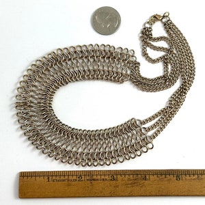 May include: A silver chain necklace with a large, layered, woven collar. The necklace is approximately 12.7 cm long.