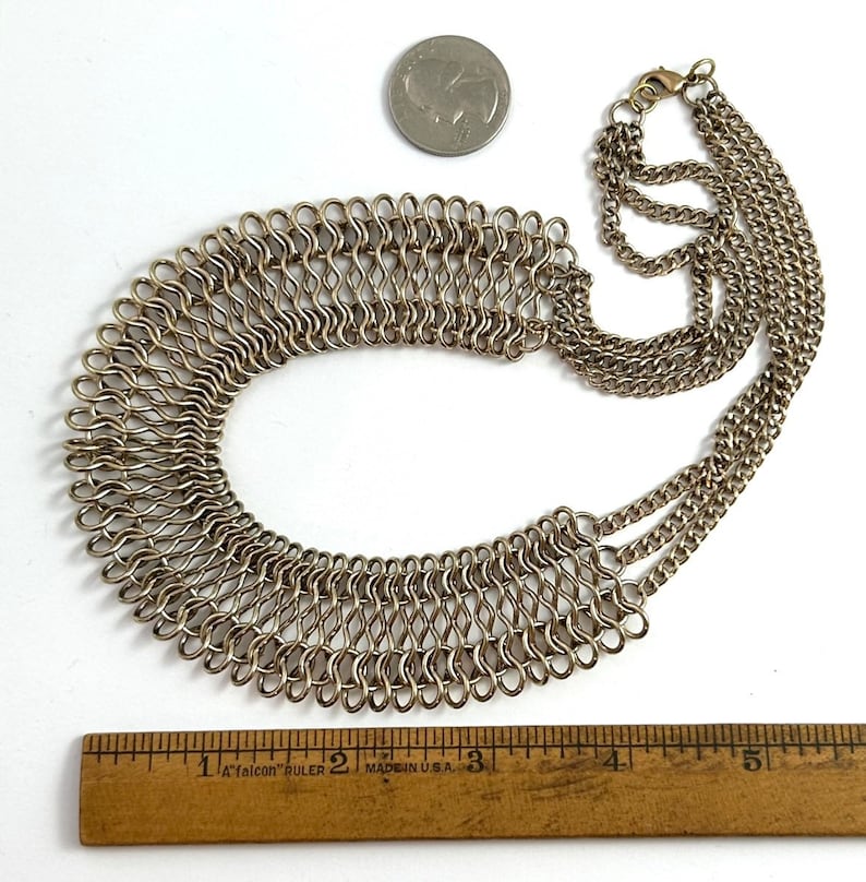 May include: A silver chain necklace with a large, layered, woven collar. The necklace is approximately 12.7 cm long.