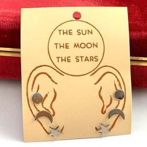 May include: A set of three silver earrings with a sun, moon, and star design. The earrings are displayed on a card with the text "The Sun The Moon The Stars".