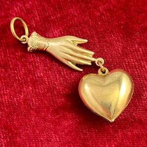 May include: A gold pendant charm featuring a hand reaching out to a heart-shaped locket. The pendant is made of gold and has a detailed design.