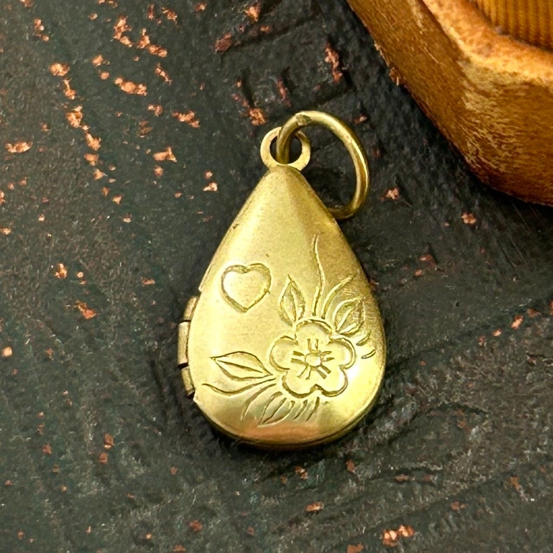 May include: A gold-toned teardrop-shaped locket with an engraved floral design and a heart. The locket has a hinged opening.