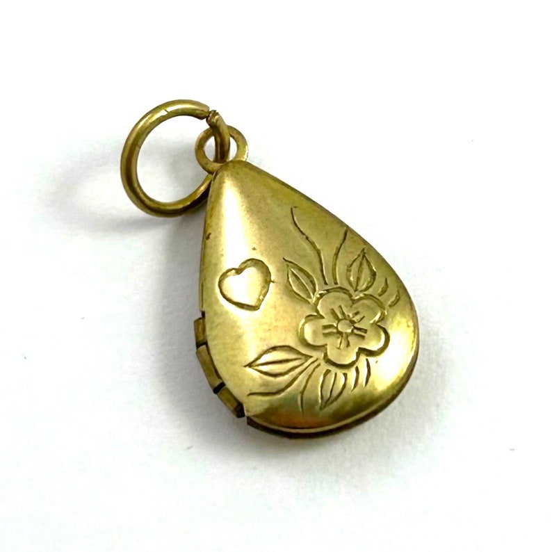 May include: A gold teardrop-shaped locket with a floral design and a heart engraved on the front. The locket has a hinged opening.