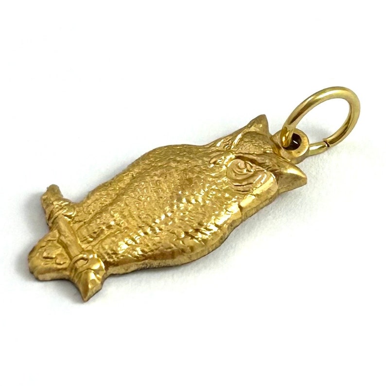 May include: A gold-colored owl charm with a detailed design. The owl is perched with its wings folded and has a round loop for attaching to a necklace or bracelet.