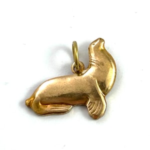 May include: A gold-toned charm in the shape of a seal. The seal is lying on its belly with its head raised and looking to the right. The charm has a loop at the top for attaching to a necklace or bracelet.
