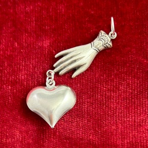 Puede incluir: Un amuleto de plata en forma de mano que se extiende hacia un amuleto de plata en forma de coraz&oacute;n.