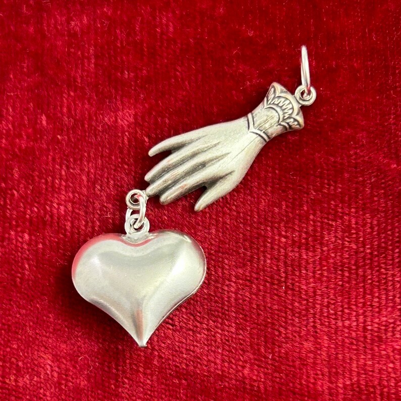 Puede incluir: Un amuleto de plata en forma de mano que se extiende hacia un amuleto de plata en forma de coraz&oacute;n.