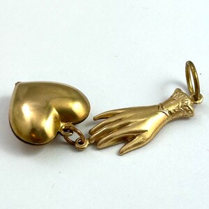 May include: A gold-toned pendant charm featuring a heart-shaped locket and a hand reaching out. The locket is open and the hand is detailed with a cuff.