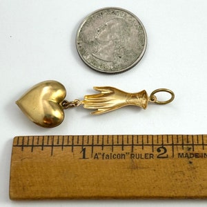 May include: A gold-toned pendant charm featuring a heart and a hand reaching out. The charm is approximately 2.5 cm long and is shown next to a ruler for size reference.