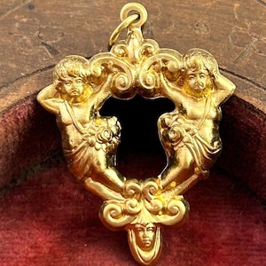 May include: A gold-colored pendant featuring two cherubic figures flanking a central void. The cherubs are adorned with floral accents and hold their heads. The pendant has a detailed, ornate design and a small loop for hanging.
