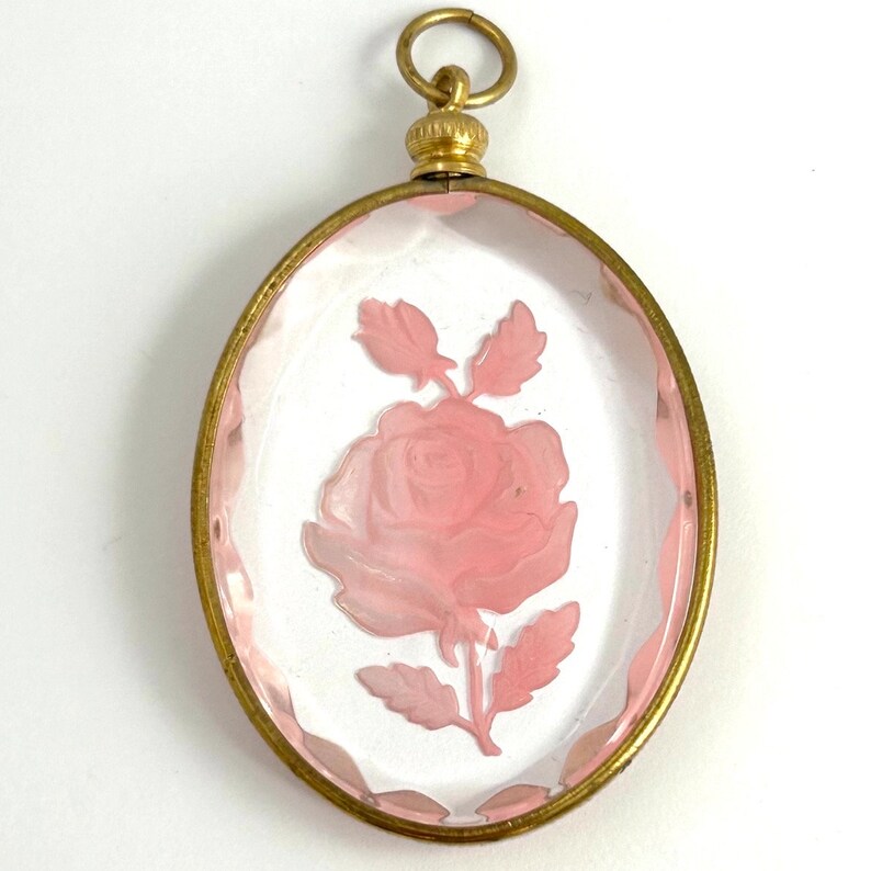 May include: A gold-toned oval locket with a clear glass front featuring a pink rose etched into the glass.