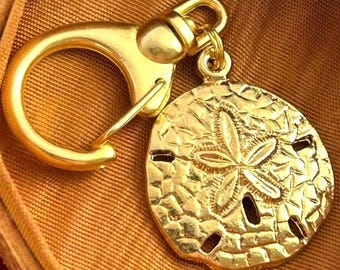 Könnte beinhalten: Goldfarbener Schlüsselanhänger mit einem detaillierten Sanddollar-Anhänger mit einem fünfzackigen Sternmuster. Der Schlüsselanhänger hat einen goldfarbenen Metallring.