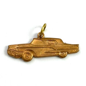Puede incluir: Un amuleto de metal de color dorado con forma de coche antiguo. El amuleto est&aacute; detallado con un perfil lateral del coche, incluyendo las ruedas, los faros y el parabrisas.