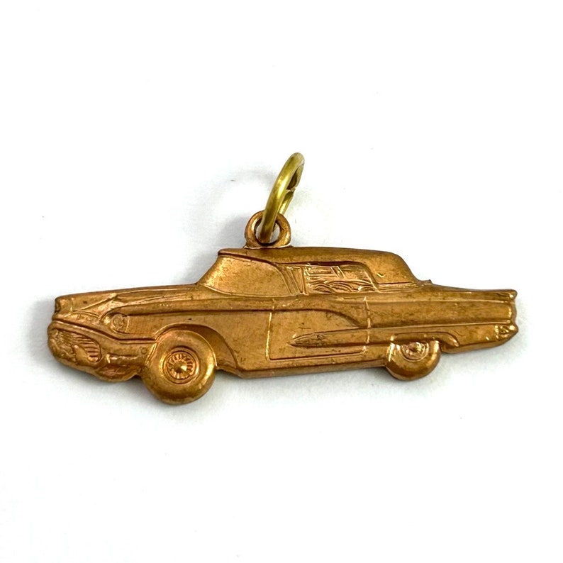 Puede incluir: Un amuleto de metal de color dorado con forma de coche antiguo. El amuleto est&aacute; detallado con un perfil lateral del coche, incluyendo las ruedas, los faros y el parabrisas.