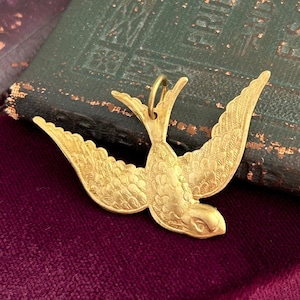 May include: A gold-toned metal pendant in the shape of a stylized bird with detailed wings. The pendant is on a dark red velvet surface.