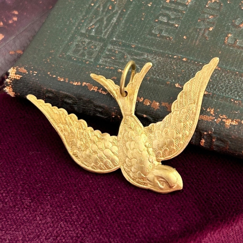 May include: A gold-toned metal pendant in the shape of a stylized bird with detailed wings. The pendant is on a dark red velvet surface.