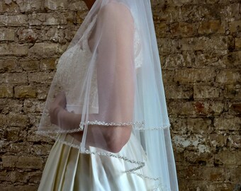May include: A single-tier tulle bridal veil with a beaded trim. The veil is worn by a person in a white satin dress.