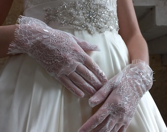 May include: A pair of white lace gloves with a delicate floral pattern. The gloves are worn by a person wearing a white wedding dress with a beaded belt.