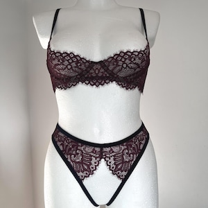 Op de afbeelding: Een tweedelige lingerie set in diep bordeauxrood, gepresenteerd op een witte mannequin. De set bevat een bh met delicate kanten details en verstelbare bandjes, en een bijpassende slip. Zwarte randen.