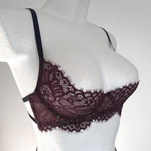 May include: A burgundy lace bra with black straps. The bra features intricate floral lace detailing and a scalloped edge. The bra is displayed on a white mannequin, showcasing its design and structure. The straps are thin and adjustable.