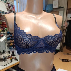 May include: A dark blue lace bra featuring a scalloped edge along the neckline and underwire cups. The garment has thin, adjustable black shoulder straps and a semi-sheer floral pattern throughout the lace fabric, displayed on a mannequin.
