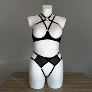 Op de afbeelding: Zwarte lingerie set op een witte mannequin. De set bevat een halter-bh met ringaccenten en een bijpassende high-waist slip met mesh details. De lingerie is gemaakt van een transparante, delicate stof.