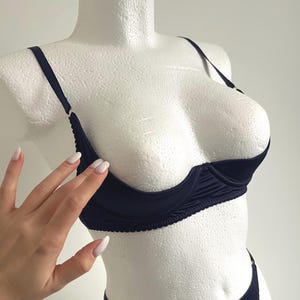 Puede incluir: Un sujetador azul marino con diseño de media copa. El sujetador tiene tirantes finos y un ribete festoneado a lo largo del borde inferior. El sujetador se muestra en un maniquí blanco.