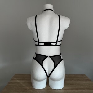 以下が含まれることがあります： マネキンに展示された黒いランジェリーセット。セットには、複数のストラップが付いたホルターネックブラと、お揃いのハイウエストショーツが含まれています。ランジェリーは、繊細なディテールが施されたシアーブラック生地で作られています。マネキンは白です。