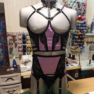 Op de afbeelding: Een lila en zwarte lingerieset bestaande uit een korset-stijl top met zwarte kanten rand, een bijpassende jarretelgordel en een zwart kunstlederen borstharnas met zilverkleurige metalen ringen en verstelbare bandjes op een paspop.