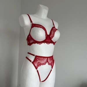 May include: A red lace lingerie set displayed on a white mannequin. The set includes a bra with delicate straps and a matching high-waisted bottom. The bra features a unique open-bust design, and the set is made of a sheer, intricate lace.