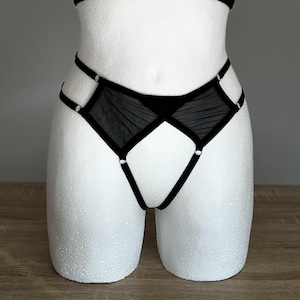 May include: Black high-waisted underwear featuring sheer mesh panels, solid black fabric sections, and thin elastic straps connected by small silver-toned metal rings. The design includes a cut-out style with a geometric arrangement of straps.