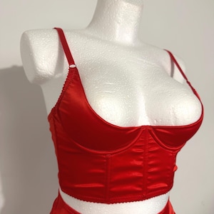 May include: A bright red satin corset-style top featuring a low-cut scoop neckline, thin adjustable shoulder straps, and vertical stitched paneling. The garment has a cropped hemline and a smooth, reflective finish, displayed on a white mannequin.