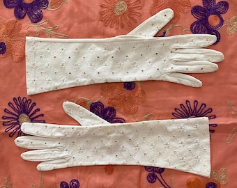 May include: A pair of white, long, embroidered gloves with a delicate floral pattern. The gloves are laid out on a peach-colored fabric with embroidered flowers in purple, orange, and green.