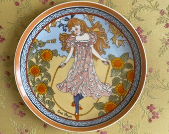 May include: A decorative ceramic plate with a colorful illustration of a girl skipping rope in a field of sunflowers. The plate has a blue and white border with a gold rim. The artist's signature, "Louis Royer", is visible at the bottom of the plate.