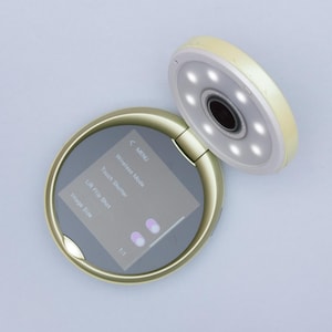 Pode incluir: Uma câmara digital redonda de cor dourada com uma secção superior rebatível que apresenta uma lente circular rodeada por seis pequenas luzes LED. A base apresenta um ecrã de menu digital com opções de texto como Wireless Mode.