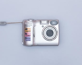 May include: Silver digital camera with a wrist strap. The camera has a lens, flash, and buttons. The camera has the following text: COOLPIX 4600, 4.0 Megapixels, 3x ZOOM, Red-eye Reduction.