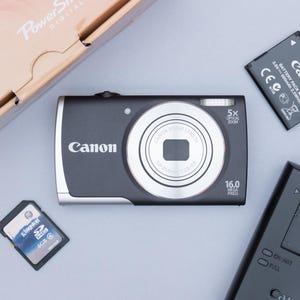 May include: A black Canon PowerShot digital camera with a silver lens, 16.0 megapixels, and 5x optical zoom. Includes a battery, a memory card, and a Canon battery charger. The camera is on a light gray surface.