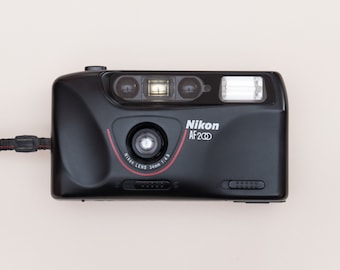 May include: A black Nikon AF200 point-and-shoot camera with a red Nikon lens ring. The camera has a built-in flash and a strap attached.