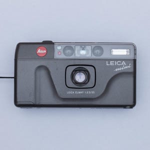 Op de afbeelding: Een grijze Leica Mini 35mm filmcamera met een zwart polsbandje. De camera heeft een rood Leica-logo en de tekst "LEICA mini" en "LEICA ELMAR 1:3.5/35" erop gedrukt. De camera is rechthoekig met afgeronde hoeken.