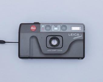 May include: A gray Leica Mini 35mm film camera with a black wrist strap. The camera has a red Leica logo and the text "LEICA mini" and "LEICA ELMAR 1:3.5/35" printed on it. The camera is rectangular with rounded edges.