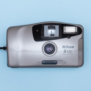 May include: Silver Nikon BF100 35mm film camera with a built-in flash and lens. The camera has a black strap attached and is set against a light blue background. The Nikon logo and model number are visible.