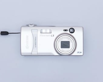 May include: A silver digital camera with a black strap. The camera has a 3x zoom lens and a 3.2 mega pixel resolution. The camera is labeled "FinePix L3" and "FUJIFILM".
