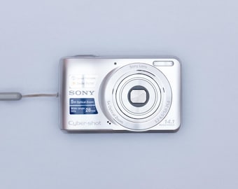 May include: Silver Sony Cyber-shot digital camera with a wrist strap. The camera features a 5x optical zoom lens and a 14.1-megapixel resolution. The wide-angle lens is 28mm. The camera is on a light blue background.