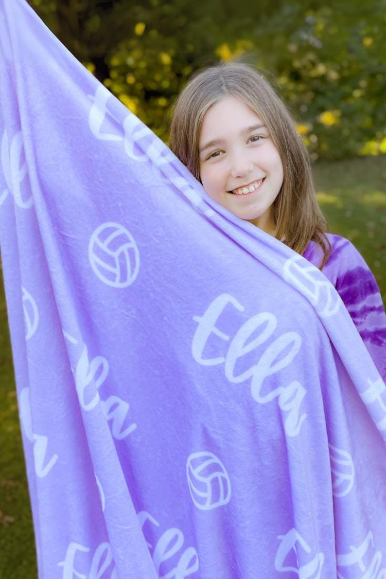 May include: A purple fleece blanket with the name "Ella" and volleyball graphics printed on it. A young girl is holding the blanket in front of her.