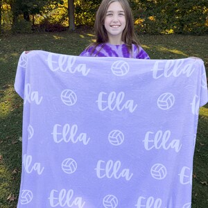 May include: A purple fleece blanket with the name "Ella" repeated throughout the design. White volleyball graphics are also printed on the blanket.