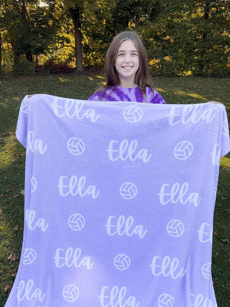 May include: A purple fleece blanket with the name "Ella" repeated throughout the design. White volleyball graphics are also printed on the blanket.