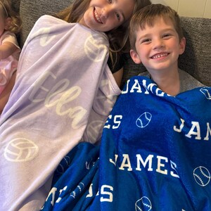 May include: Two children are sitting on a couch, each with a personalized fleece blanket. The girl's blanket is lavender with the name "Ella" and white volleyball designs. The boy's blanket is blue with the name "James" and white baseball designs.
